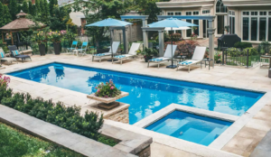 Rectangular fiberglass pool with built-in spa, stone paver deck, lounge chairs, and pergola in a landscaped backyard