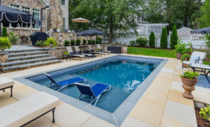 Fiberglass pool with built-in tanning ledge, submerged lounge chairs, decorative tile, and upscale stone patio setting