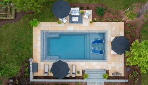 Aerial view of an inground fiberglass pool with sun shelf, surrounding paver patio, patio umbrellas, and lush backyard landscaping