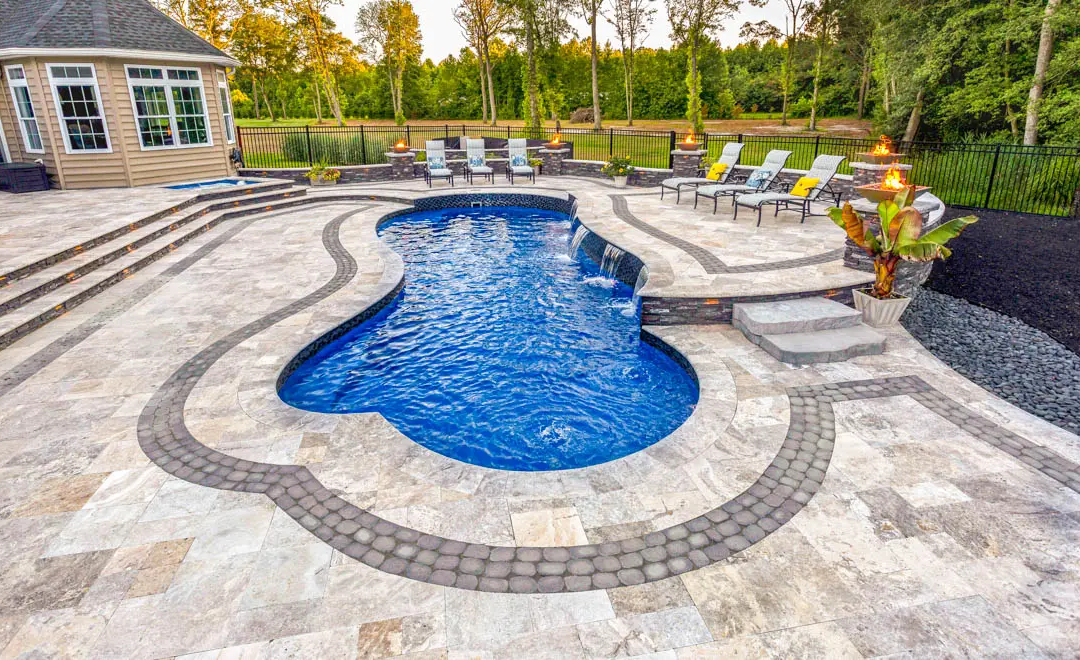 Freeform fiberglass pool with waterfall, decorative travertine paver deck, fire features, and spa in a resort-style backyard