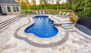 Freeform fiberglass pool with waterfall, decorative travertine paver deck, fire features, and spa in a resort-style backyard